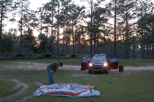 A man bends over, exhausted, over a not-yet-set up tent in a grassy field crossed with tire tracks. Behind him, the sun sets through sparse trees and over what might be drainage openings in a retaining wall while a woman unloads a car.