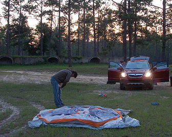Summary graphic: A man bends over, exhausted, over a not-yet-set up tent in a grassy field crossed with tire tracks. Behind him, the sun sets through sparse trees and over what might be drainage openings in a retaining wall while a woman unloads a car.