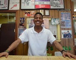 A young Black man stands behind a counter, surrounded by menu boards and family photos. He is smiling and spreading his hands out across the counter.