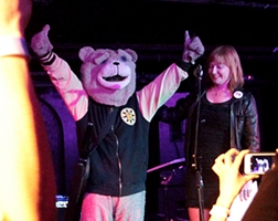 Summary graphic: Keytar Bear, wearing his full-body bear costume, stands on a stage before fans with his hands raised in a “victory” pose.