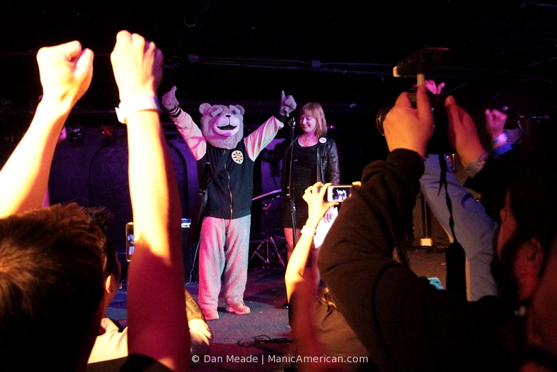 Keytar Bear, wearing his full-body bear costume, stands on a stage before fans with his hands raised in a “victory” pose.