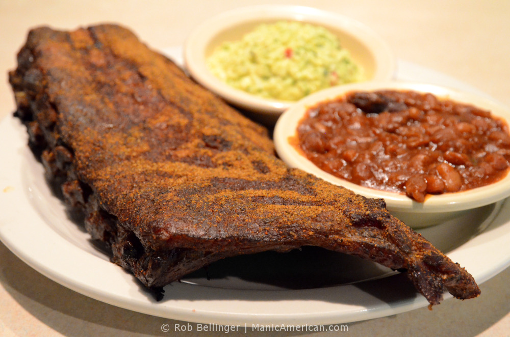 A plate of smoked pork ribs covered with dry rub, accompanied by side dishes of beans and slaw
