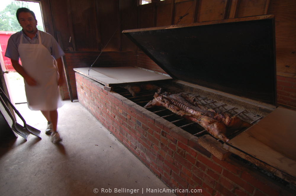 A hog smoking over hickory coals as a worker walks by