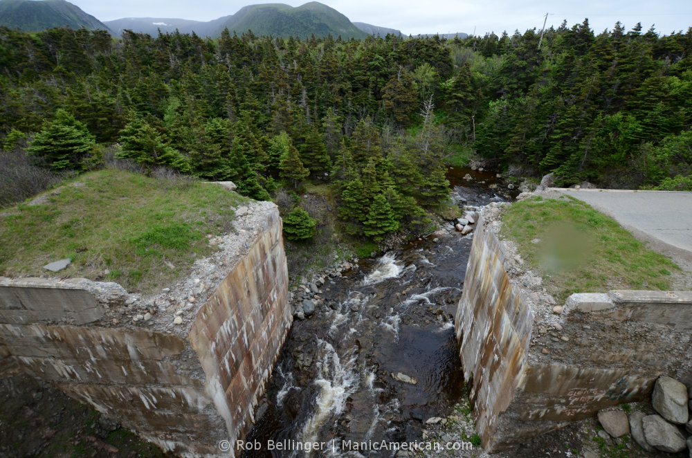 A brook babbles through the rust-stained abutments of a former brige, with pine trees and mountains in the background. Southwest Newfoundland.