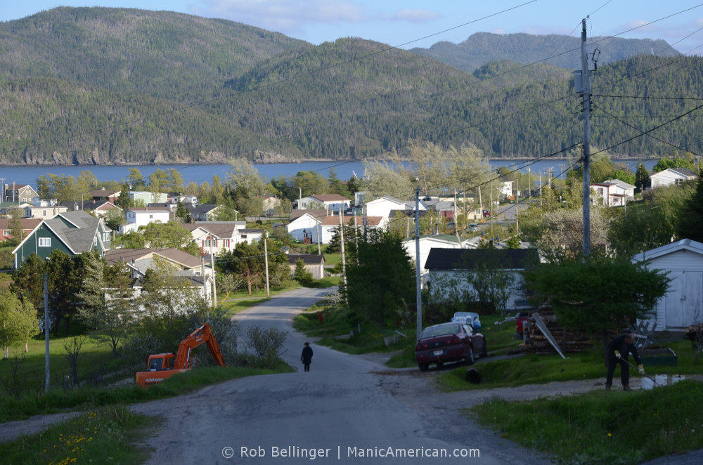 A lone, silhouetted figure walks down a winding road lined by houses, with a fjord and tree-lined mountains in the background. Bonne Bay, Newfoundland.