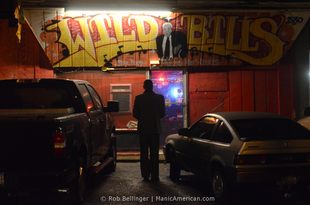 A man warming up on his saxophone stands in silhouette between two cars in front of a bar. Above is a hand-painted sign announcing that this bar is WILD BILL’S.