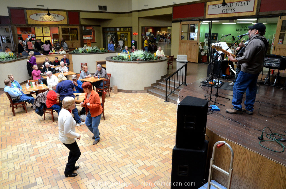 Two older women dance to the music of a band playing in front of stores in a mall while others eat lunch in the background