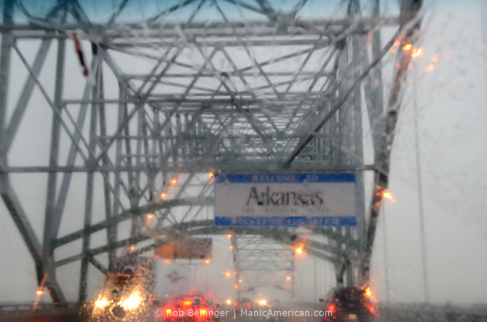 View through the windshield of a car, in heavy rain and gray skies, at the Welcome to Arkansas sign in the middle of the Hernando de Soto Bridge over the Mississippi River