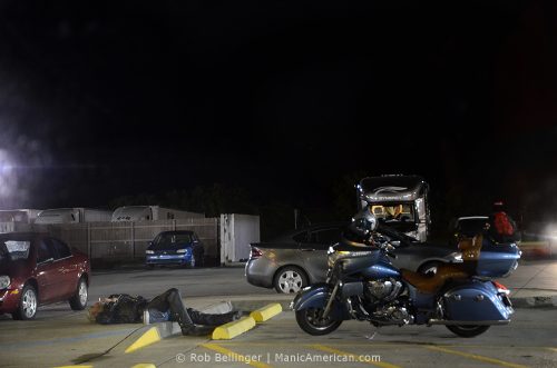 At a rural truck stop in the middle of the night, a middle-aged biker lies on the pavement, inhaling through a lit cigarette near his blue motorcyle