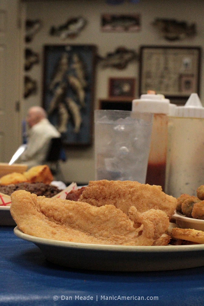 A plate of catfish in the foreground, a man before a wall of fish in the back.