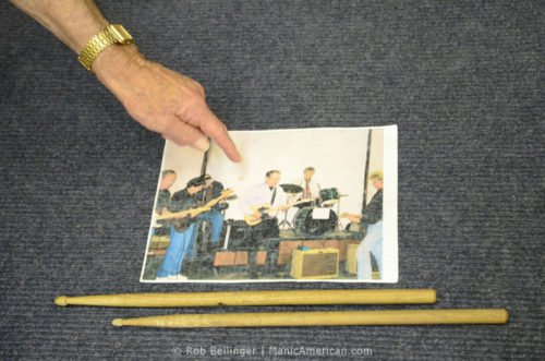 A hand, wearing a gold watch, points to a photo of musicians in the recording studio. Two drumsticks sit next to the photo.