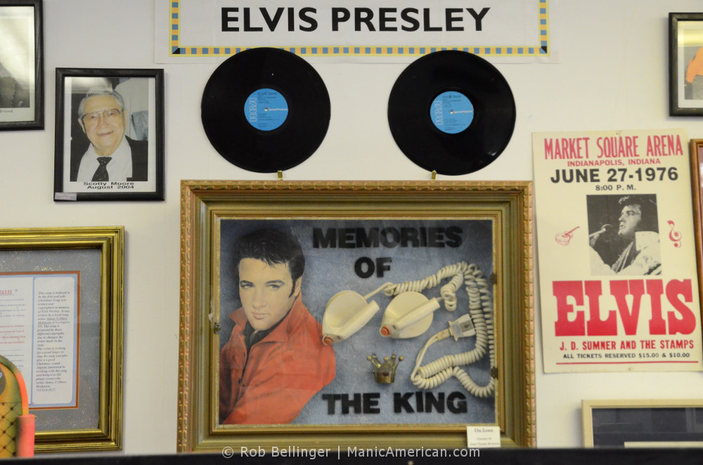 A wall of Elvis Presley memorabilia, such as records and posters, prominently features two defibrillator paddles in a glass case