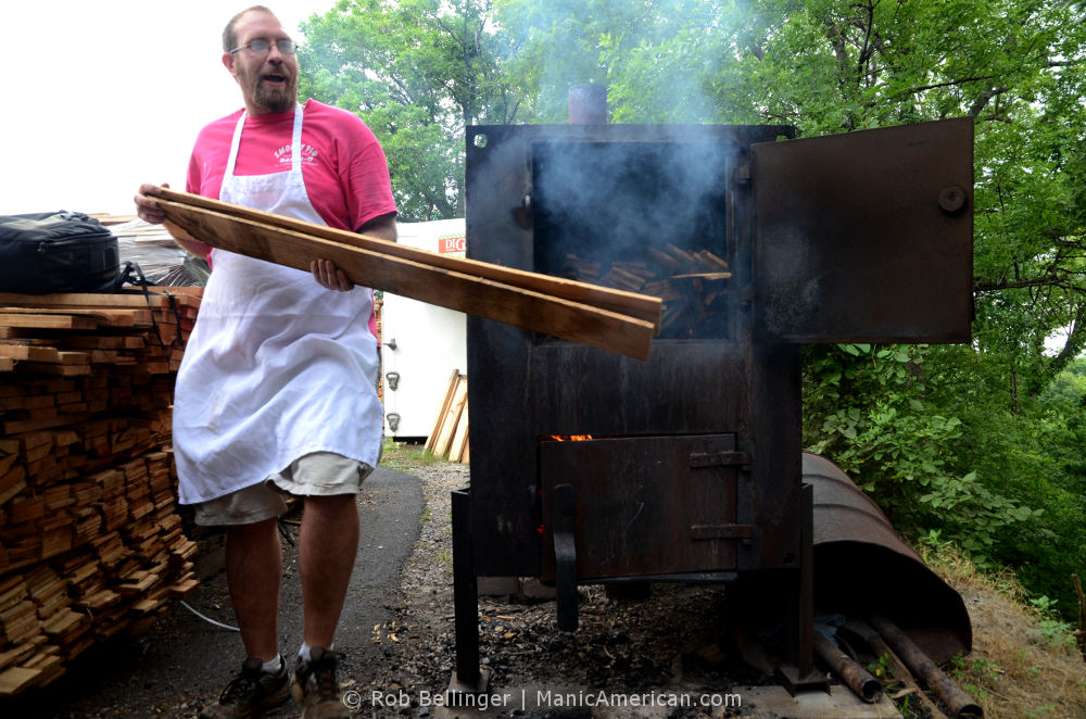 A man in an apron prepares to place two flat hickory boards into a steel furnace