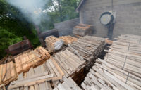 Outside a cinder block building, pallets of hickory boards sit while smoke billows from a steel cauldron in the background