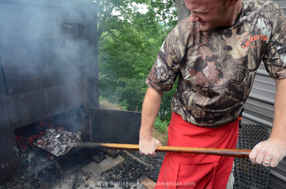 A man in a camoflauge t-shirt carefully extracts a shovelful of burning hickory coals from the base of a steel furnace, part of the process for making Kentucky barbecue.