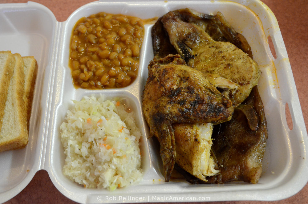 A styrofoam train containing Kentucky barbecue meats, including a chicken thigh, accompanied by beans, white cole slaw, and toast.