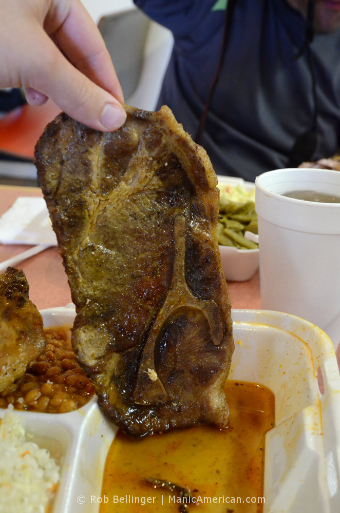 A hand lifts a slice of cooked Kentucky barbecue pork shoulder from a Styrofoam tray, revealing the juices it was dipped in