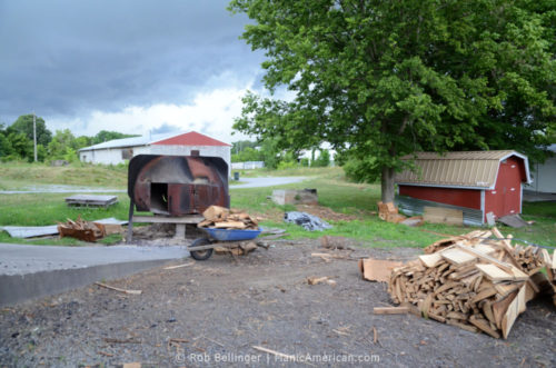 A backyard lot with a pile of hickory logs, a wheelbarrow full of logs, and a steel furnace. In the distance, dark rain clouds are forming.