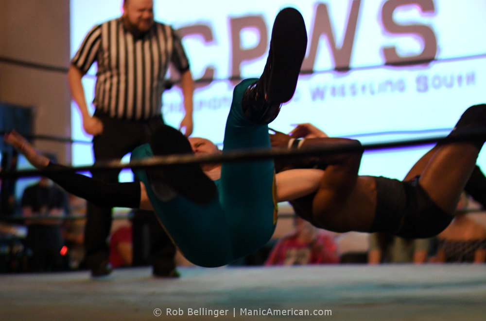 Two wrestlers fall to the mat, amrs locked, while the referee watches