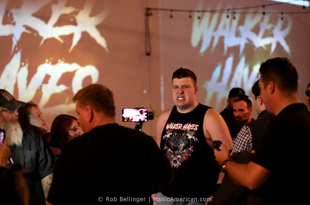 Wrestler Walker Hayes approaches the ring surrounded by fans taking pictures and video. He wears a shirt showing his name in heavy metal font and a wolf surrounded by bloody bones.