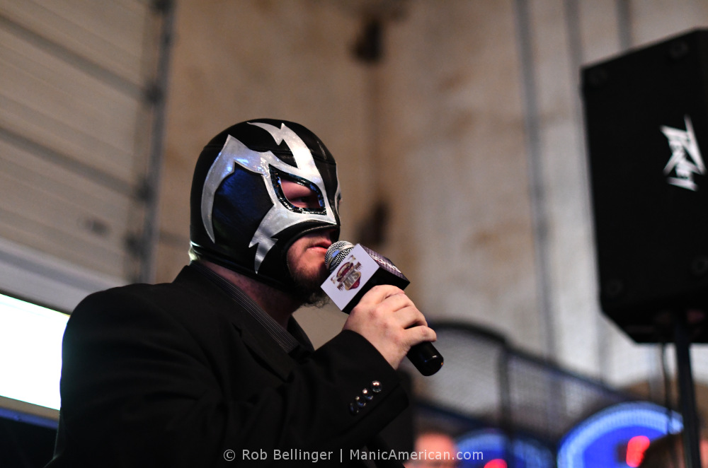 A man wearing a full-face lucha libre mask uses a microphone to announce the fight