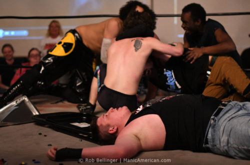 Three wrestlers help Sam Armstrong up after Walker Hayes is defeated. Hayes lies on the mat surrounded by Legos and a folding chair.