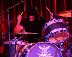 Marky Ramone plays the drums in front of the red curtain backdrop of The Bell House.