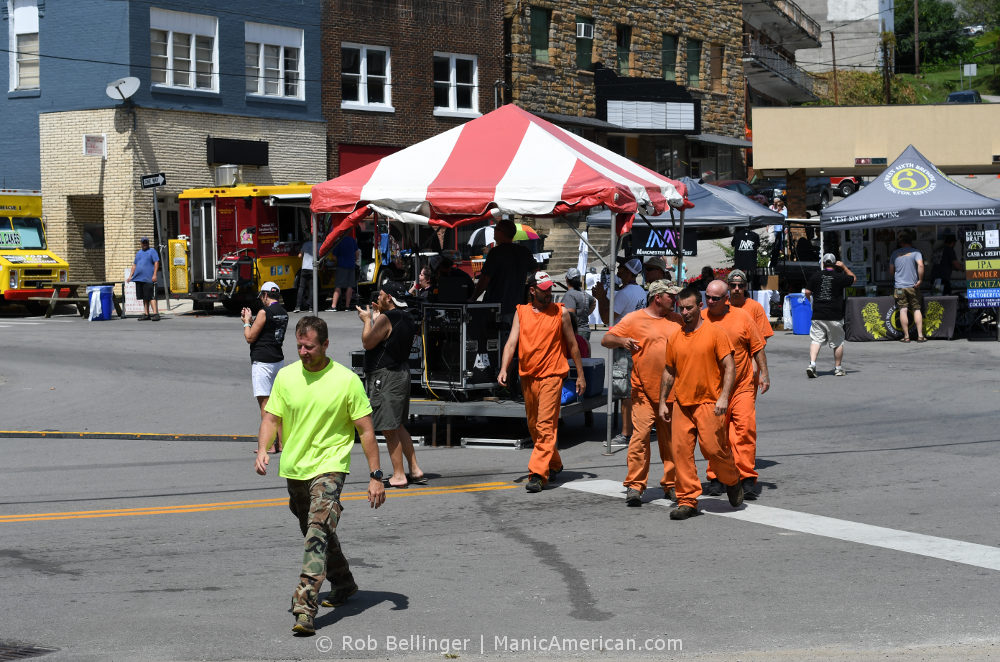 A man leads a chain gang of five prisoners in orange jump suits through a small downtown