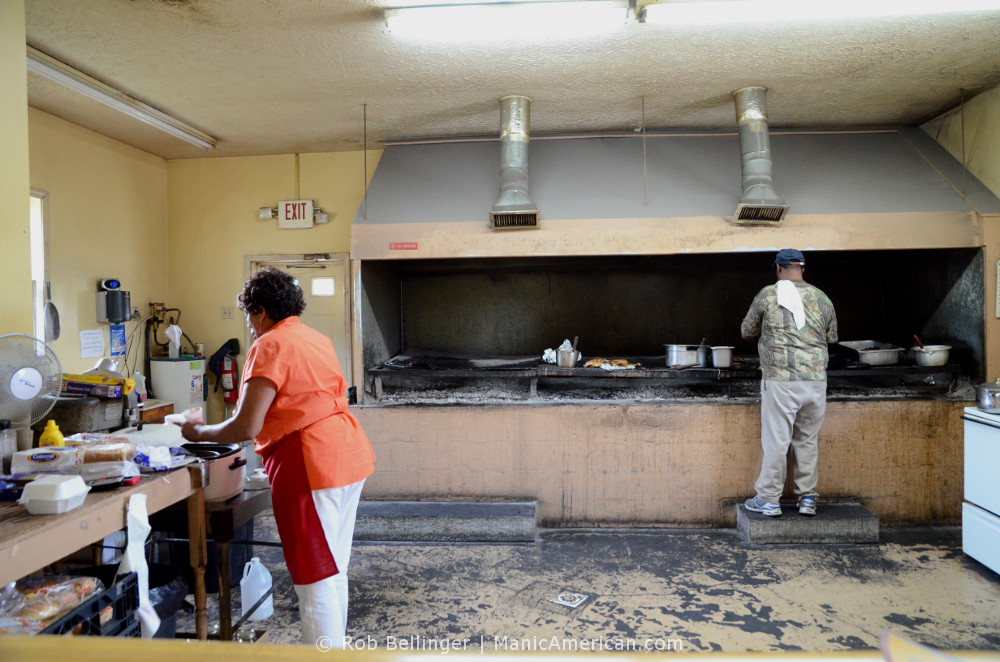 The kitchen of a Kentucky barbecue restarant, with a man tending the grill and a woman scooping beans from a slow cooker.