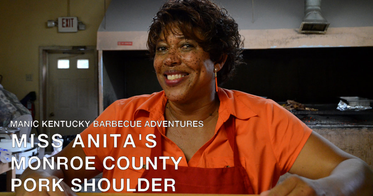 A woman in an apron stands in front of a large barbecue grill