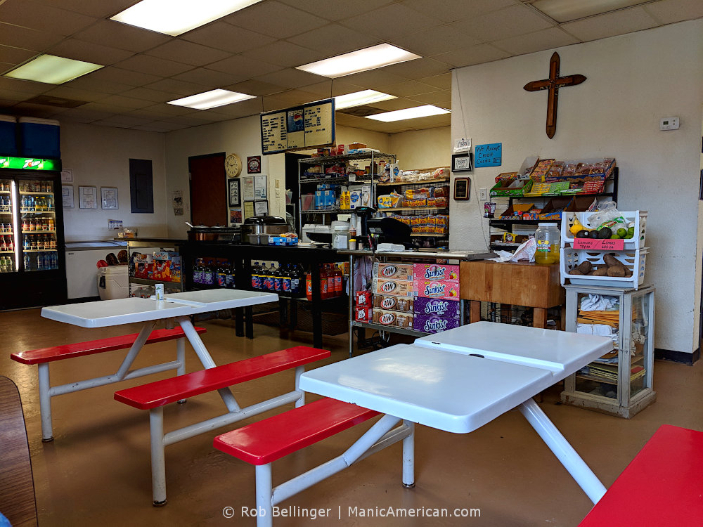 The interior of a small convenience store and restaurant with a wooden cross on the wall, a serving counter, and two metal tables with benches