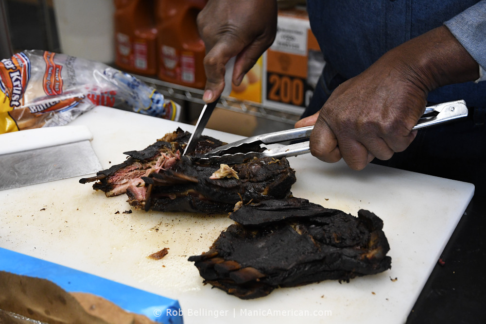 A man's hands use tongs and a knife to break apart two racks of smoked, blackened lamb ribs