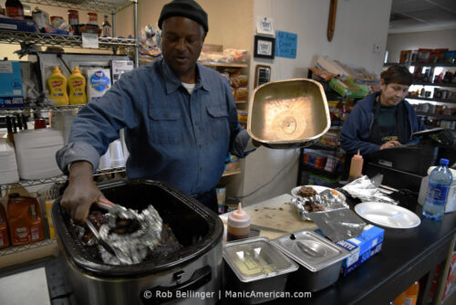 A Black man wearing a denim, button down shirt and winter cap retrieves meats from a slow-cooker at the counter of his restaurant while a woman tallies up an order on the cash register