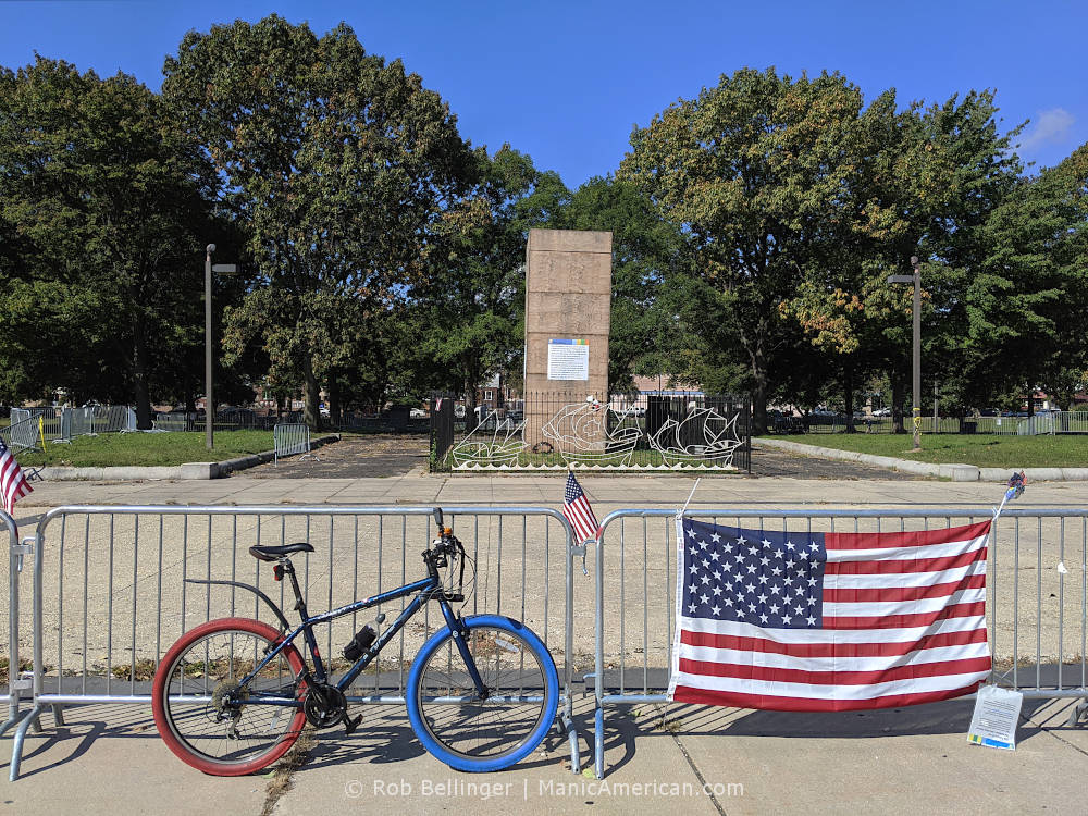 a bike with red and blue tires parked next to a fence with an american flag. in the background, a statue is in a wooden box.