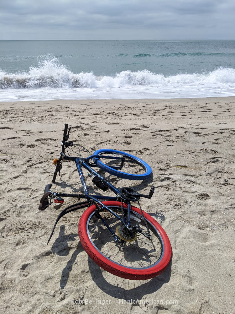 A bike with one red and one blue tire lying on rockaway beach as a wave breaks in the background