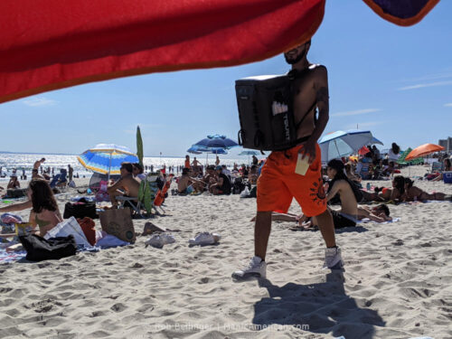 a shirtless man selling alcoholic beverages on rockaway beach using a cooler backpack mounted to his chest. his face is obscured by a beach umbrella