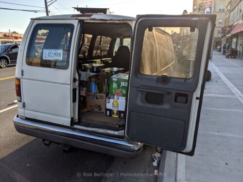 a white van with rear door open revealving cases of beer, with a fake license plate that reads ASSMAN