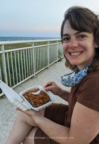a young woman eats a plate of lo mein noodles on the rockaway beach boardwalk