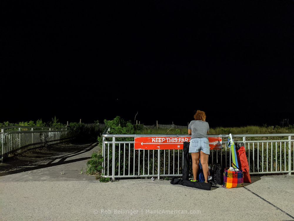 a woman looks at the ocean from the rockaway beach boardwalk, with the now-famous NYC Parks social distancing sign showing how far 6 feet is just in front of her