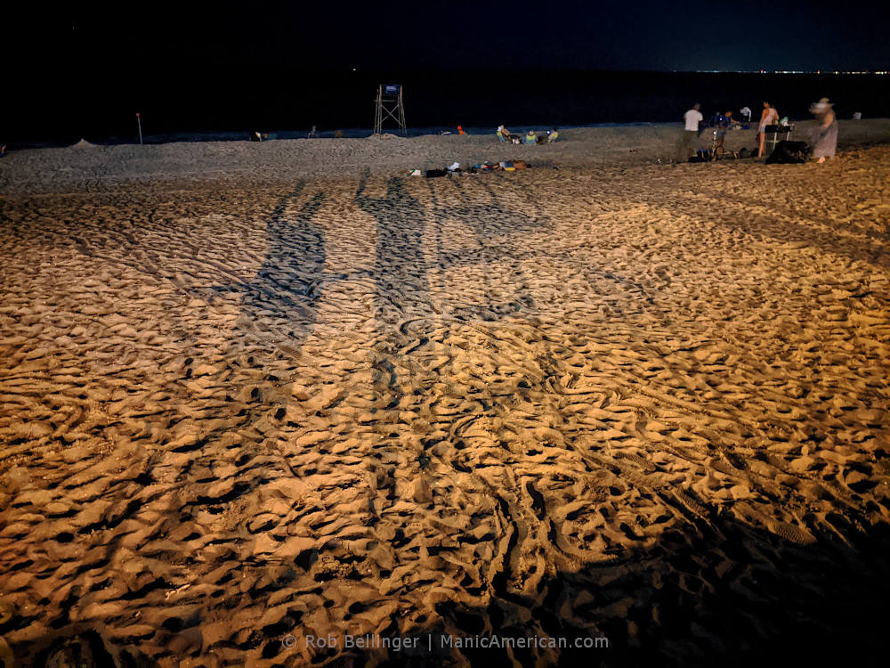 on the rockaway beach sand, two long, shadowed figures wave at the camera