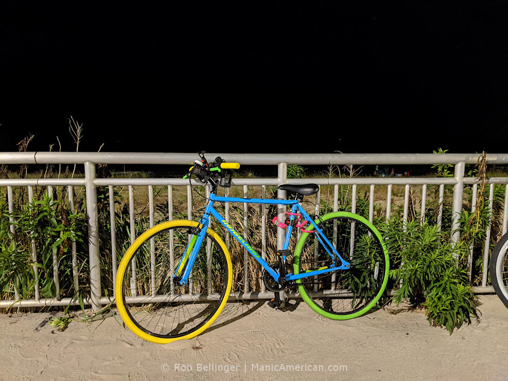 a brightly colored bike chained to the rockaway beach boardwalk at night. the bike is a Kent Ridgeland, a $99 fixed-gear bike from Walmart, which sold out at the very beginning of the pandemic bike boom.