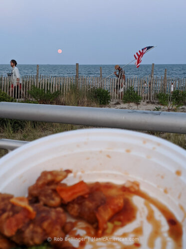 a full moon rises over the ocean, while a man carries an american flag on the rockaway beach boardwalk, viewed from behind a takeout container of orange beef