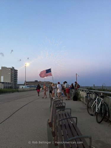 fireworks exploding over the rockaway beach boardwalk while a group of people display an american flag