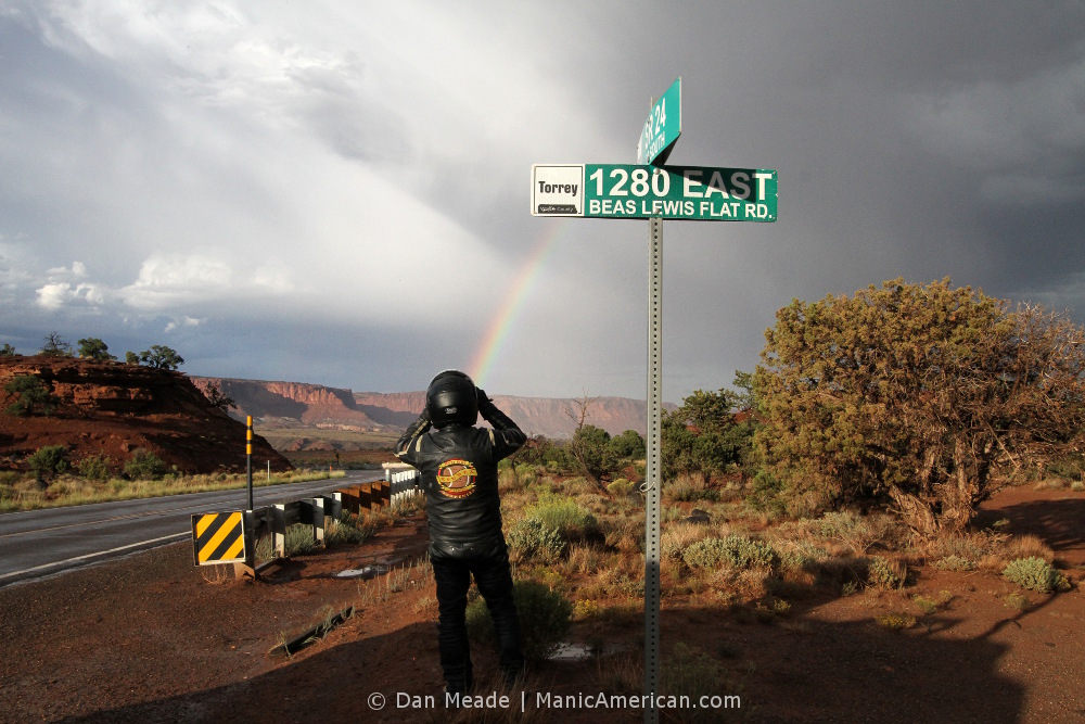 A black-leather clad motorcycle rider takes a photo of a rainbow arcing over a landscape of arid buttes and red rocks near the outskirts of Torrey, UT.