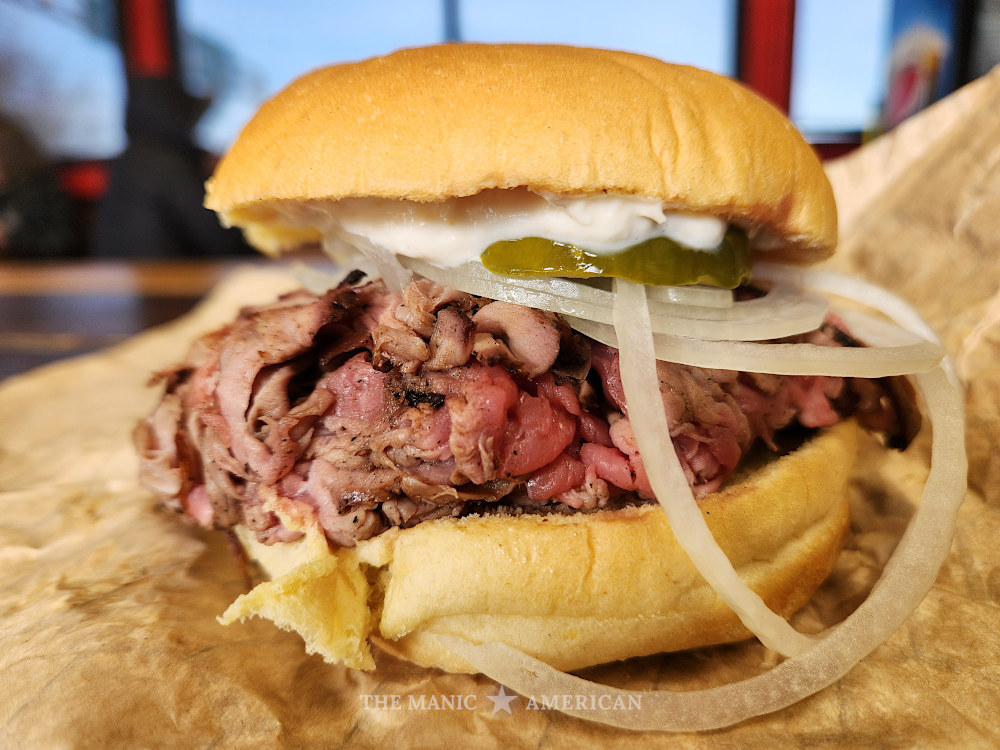 A fully dressed pit beef sandwich: medium rare beef on a potato roll, topped with wide white onions that encircle the sandwich. White sauce and green pickles are visible inside the sandwich.