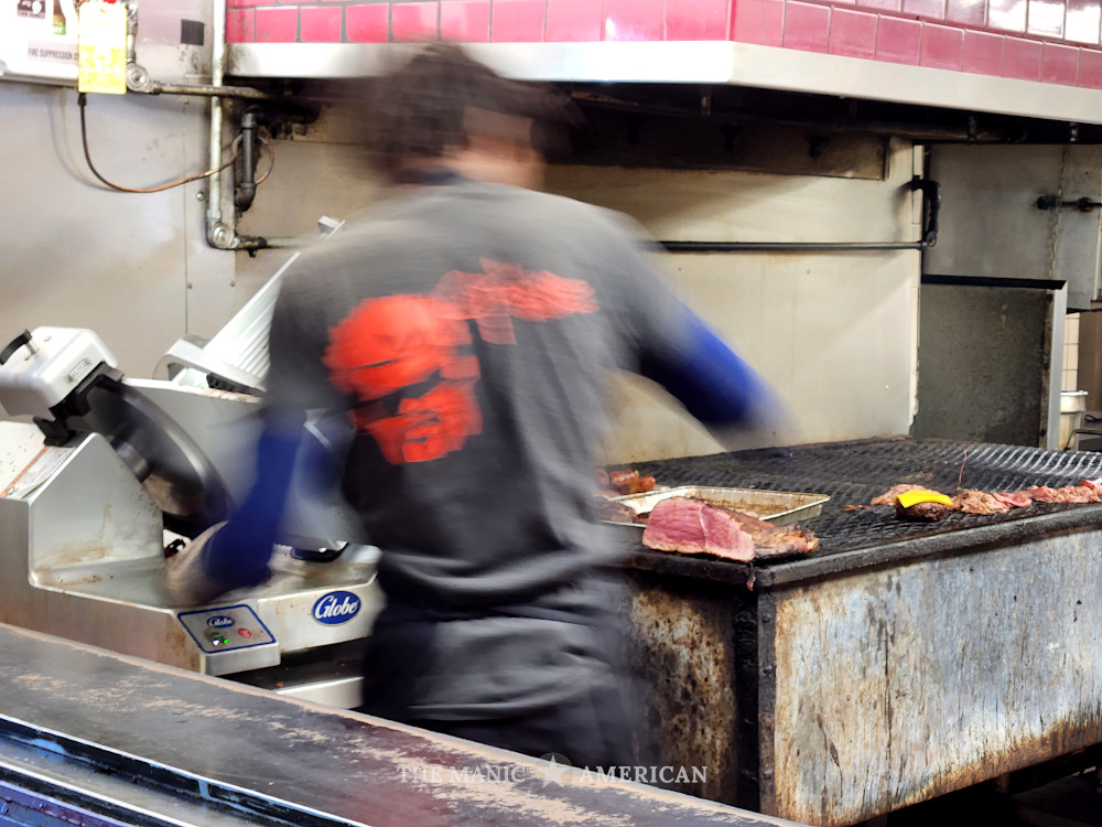 A blurred male figure operates a stainless steel meet slicer while meats that are being kept warm or grilled sit on a large charcoal grill to the man's right.