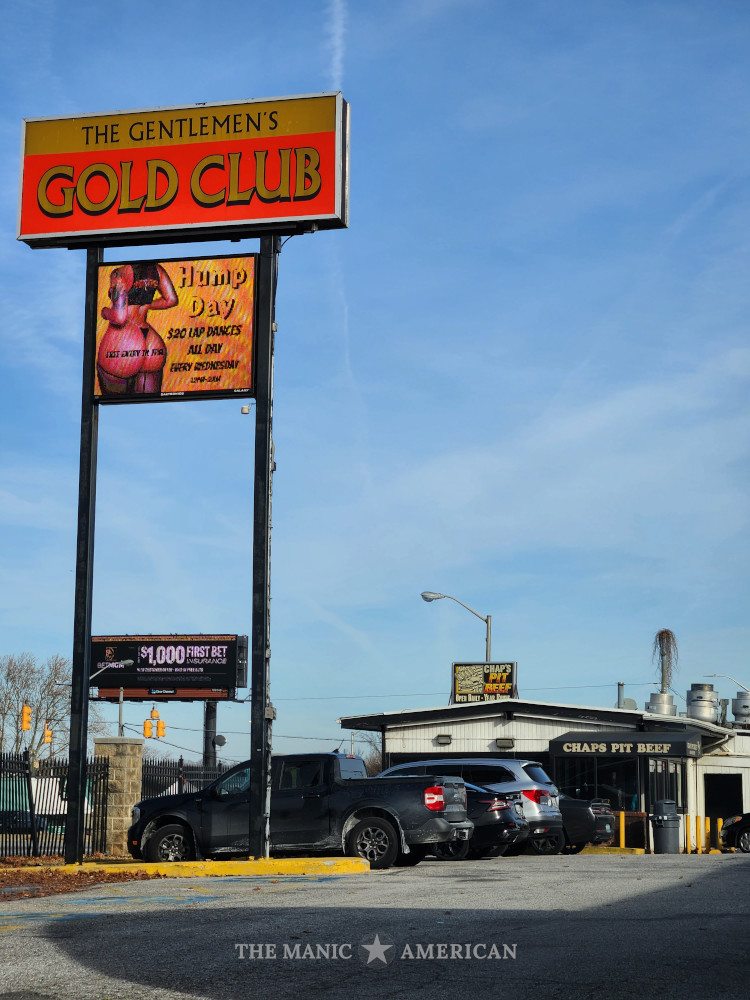 The sign for the Gentlemen's Gold Club towers above the single-story Chaps Pit Beef Restaurant. Text on a large monitor mounted below the sign advertises Hump Day, with $20 lap dances, and a depiction of a woman wearing a thong.