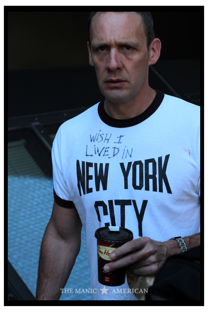 A man walking and looking toward the camera in a white t-shirt that originally read, “NEW YORK CITY” until someone wrote on it. The shirt now reads, “WISH I LIVED IN NEW YORK CITY.”